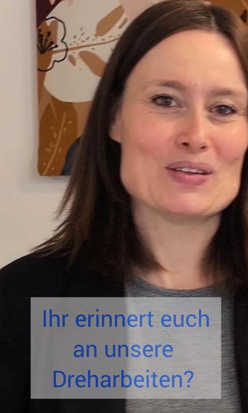 Eine dunkelhaarige Frau in einem grauen Hemd blickt direkt in die Kamera und fragt: „Ihr erinnert euch an unsere Dreharbeiten?“